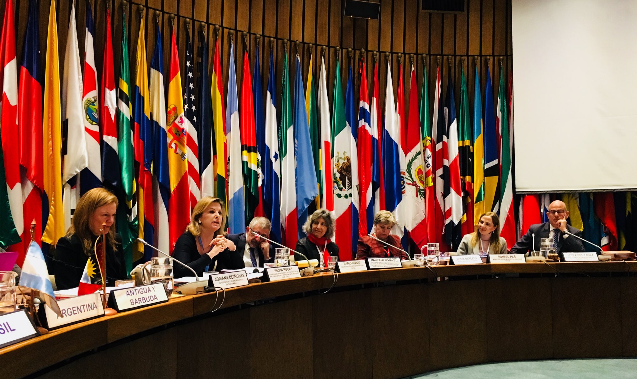 Mujeres y hombres de la ONU sentados frente a un escritorio con banderas de américa latina detrás